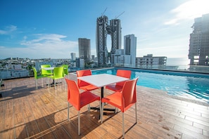 A rooftop pool area with a wooden deck, a white table, and orange chairs.