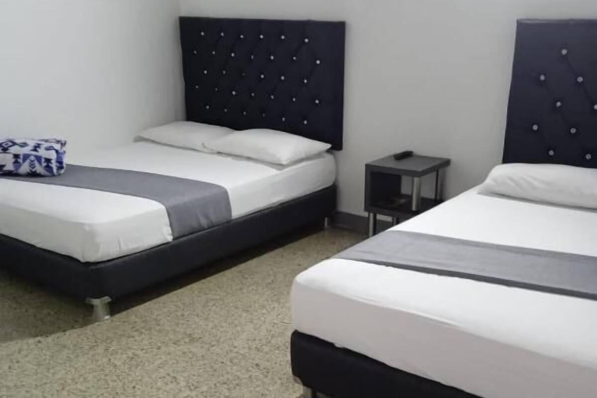 Een hotelkamer met twee bedden, elk met een donker hoofdeinde en wit beddengoed. Aan een kant staat een klein nachtkastje met een lamp.