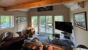 A spacious living room with a high ceiling, wooden beams, a large flat-screen TV, comfortable seating, and large windows offering a view of the outdoors.