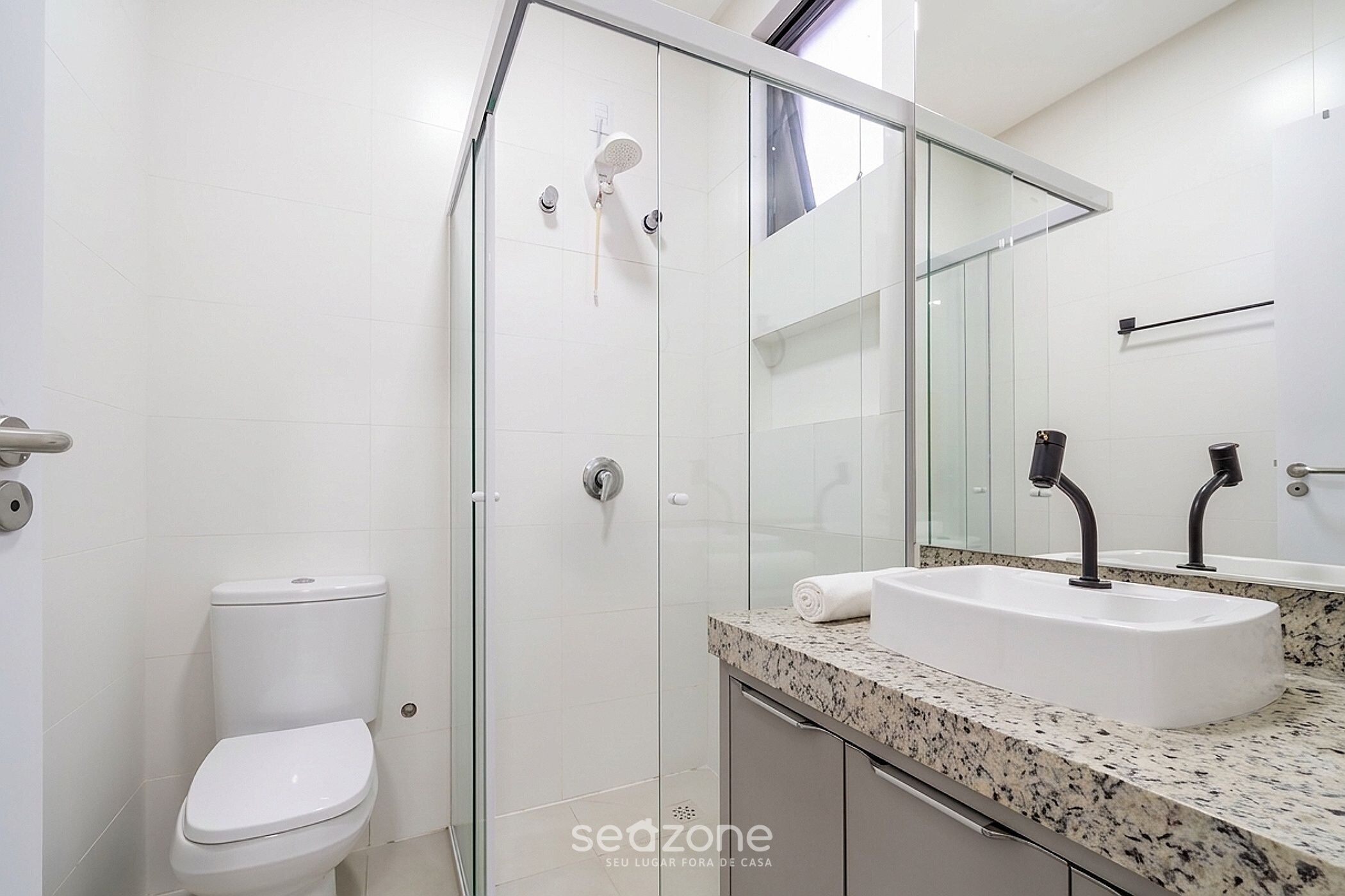 Un baño moderno con ducha envuelta en vidrio, inodoro blanco y lavamanos sobre encimera de granito.