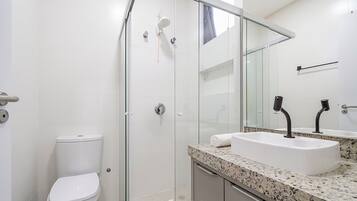 Un baño moderno con ducha envuelta en vidrio, inodoro blanco y lavamanos sobre encimera de granito.