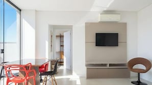 Una sala de estar moderna con un televisor de pantalla plana montado en la pared, un comedor con una mesa y sillas de vidrio, y un ventanal que proporciona luz natural.