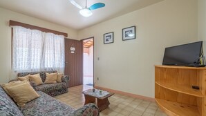A living room with a sofa, a coffee table, a television on a wooden stand, and a ceiling fan. - Casa a 4 min a pé da praia em Porto Belo BRR0097 (Porto Belo)