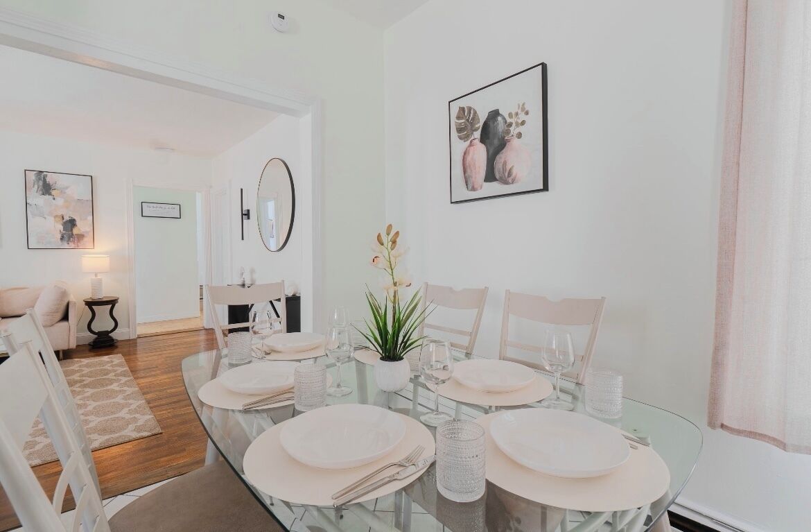 A dining area with a glass table set for four, surrounded by white chairs. There is a small potted plant on the table, and a framed artwork above it.