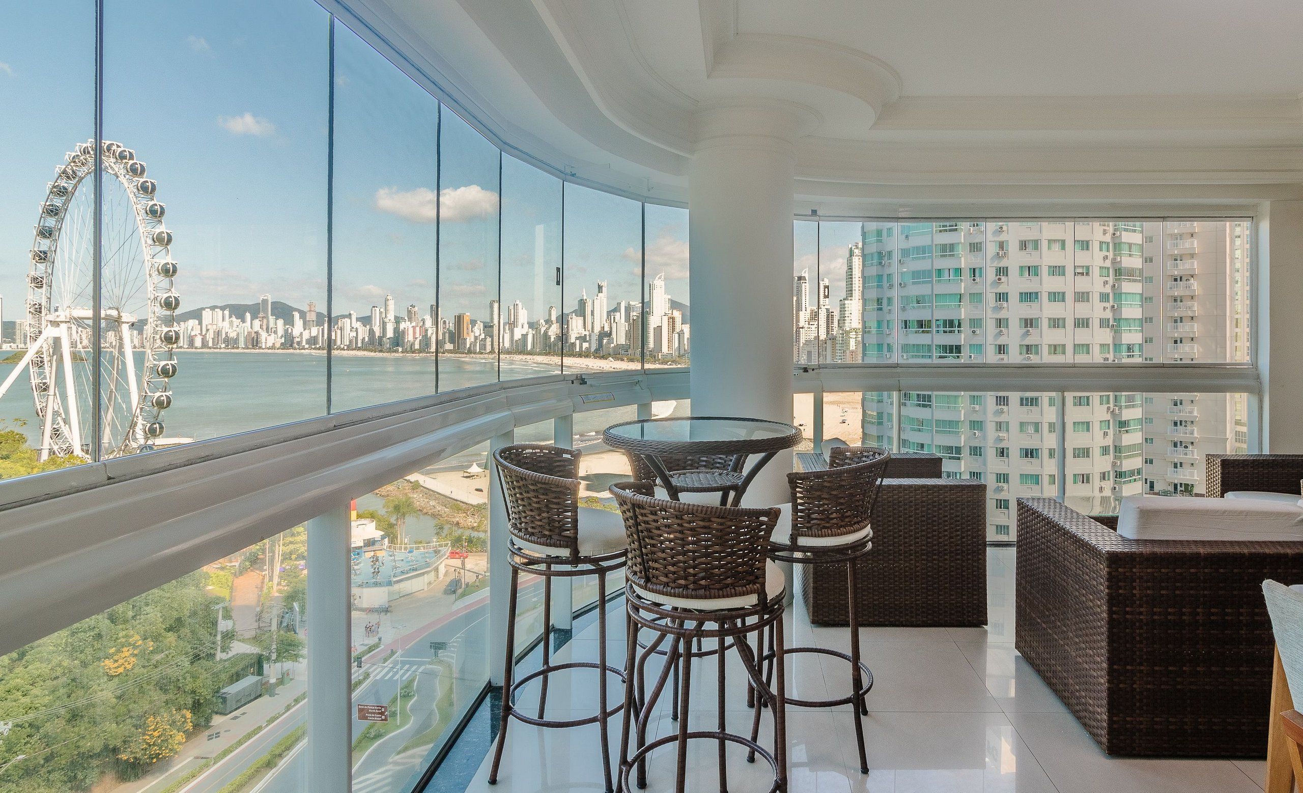 A balcony with wicker furniture and a glass railing overlooking a cityscape and a Ferris wheel.