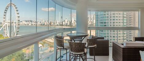 A balcony with wicker furniture and a glass railing overlooking a cityscape and a Ferris wheel.