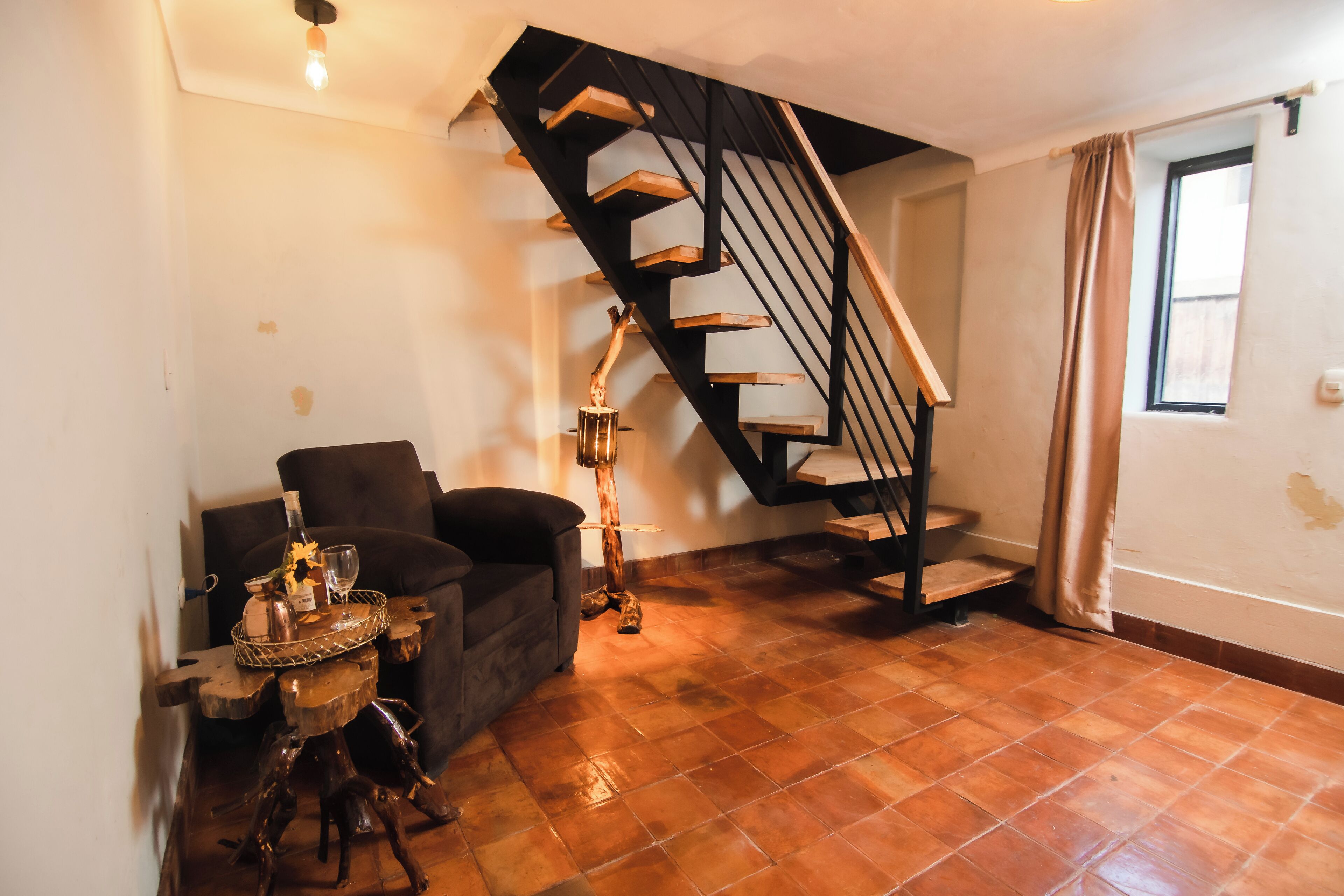 A living room with a wooden staircase, a sofa, a small table with a bottle and glass, and a floor lamp.