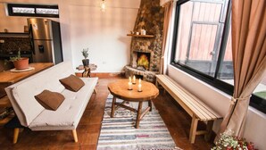 A cozy living area with a sofa, a round wooden table with candles, a stone fireplace, and a large window with curtains. - Family friendly 5-bedroom home in the Sacred Valley (Calca)