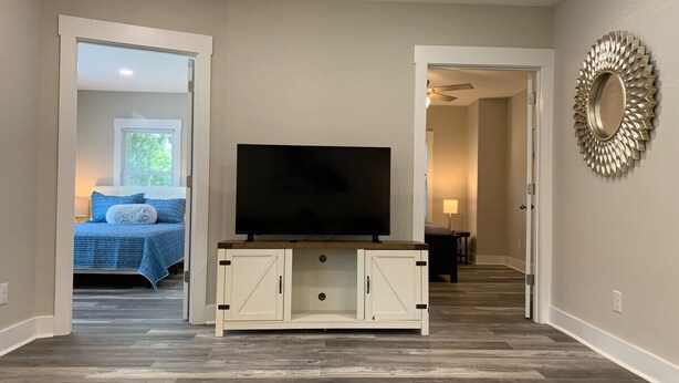 A living room with a flat-screen TV on a white cabinet, a hallway leading to a bedroom with a blue bedspread, and a decorative mirror on the wall. - Three Bedroom Downtown Cottage (Lincolnton)