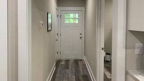 A narrow hallway with a white door at the end, a small trash bin, and a light switch on the wall. - Three Bedroom Downtown Cottage (Lincolnton)