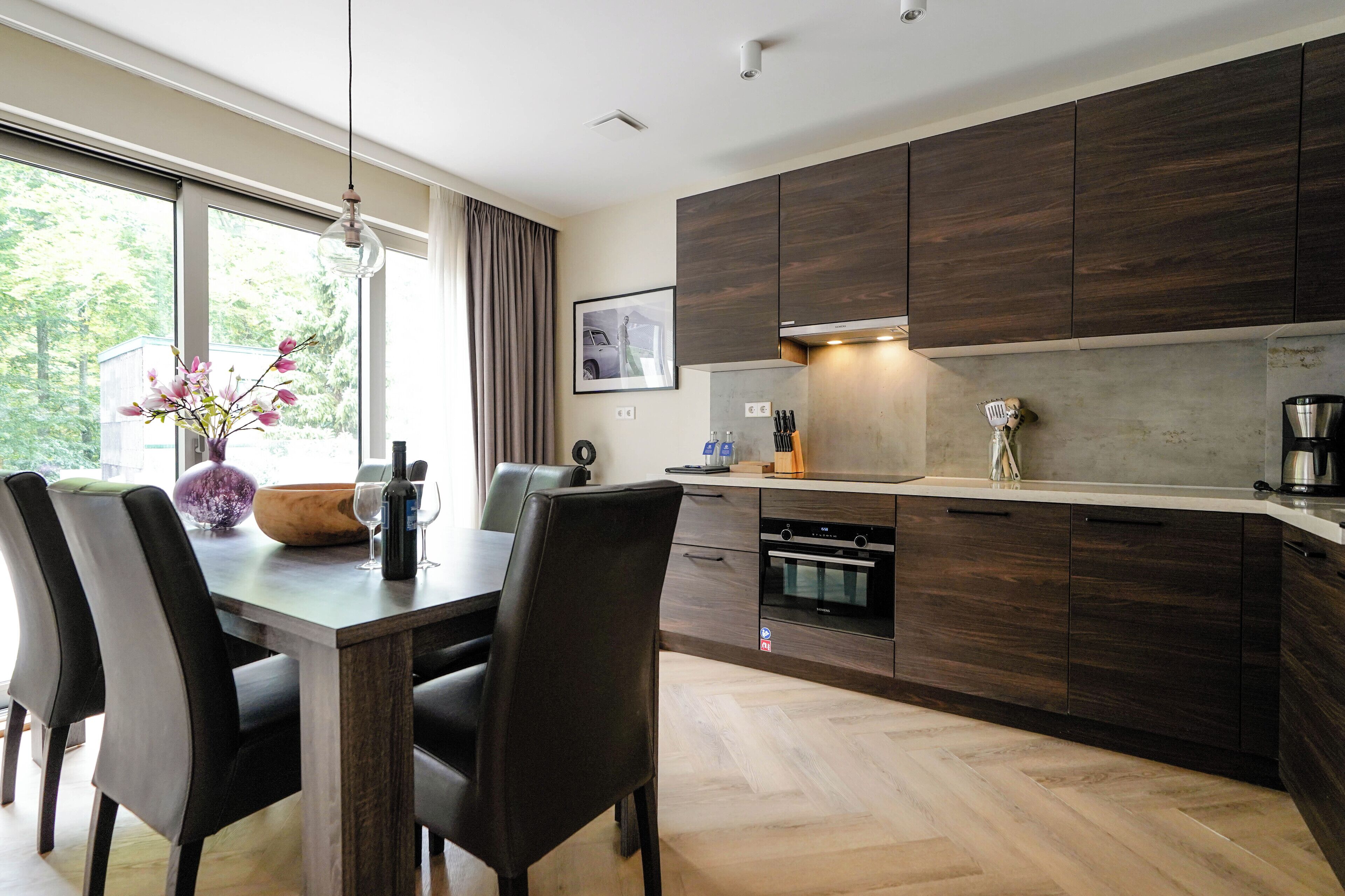 Una cocina moderna con armarios de madera oscura, un comedor con mesa y sillas de madera, y un ventanal con vistas a la vegetación.