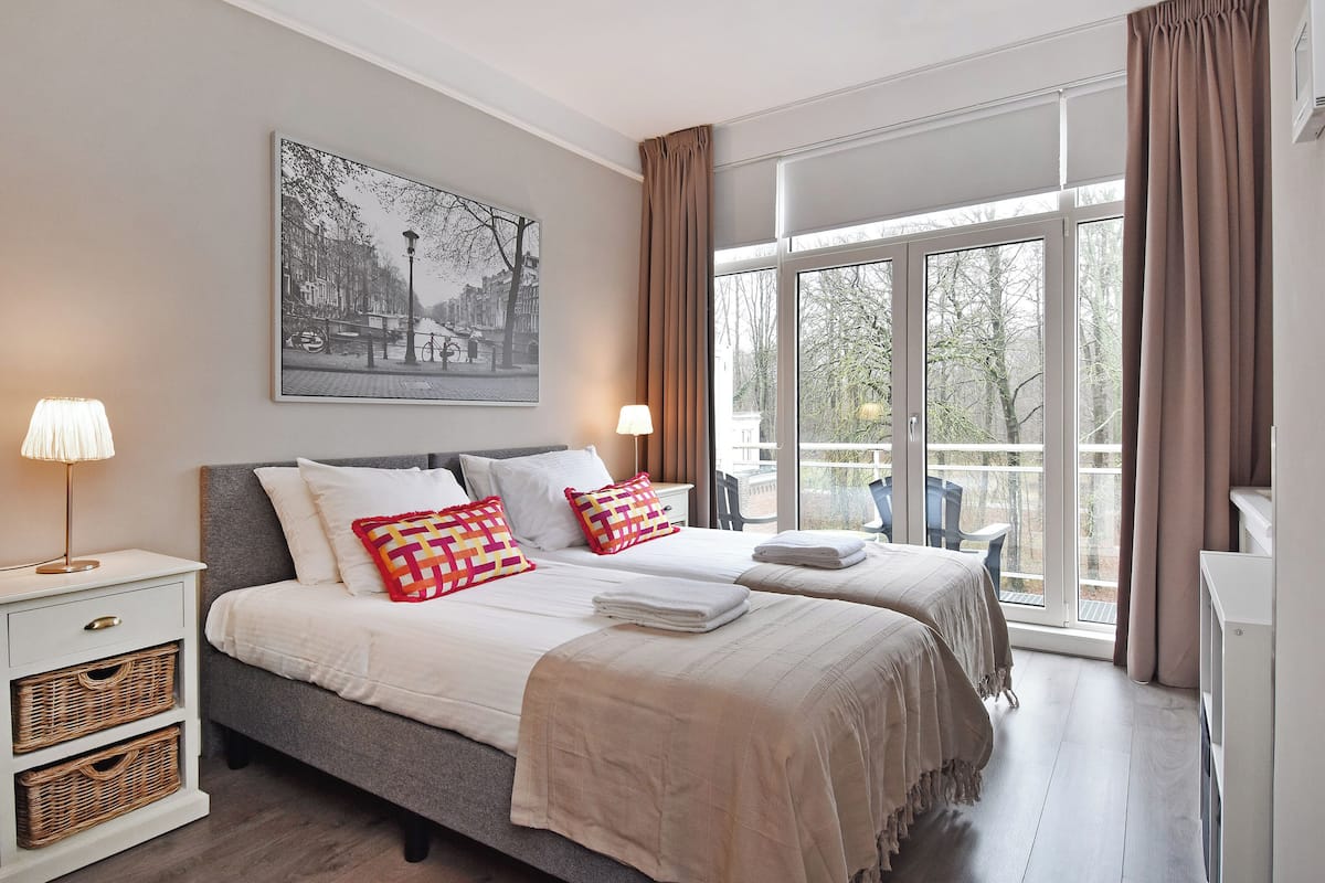 A bedroom with two beds, a nightstand, wicker baskets, a lamp, and a large window with curtains.