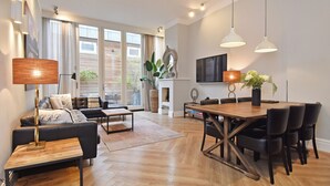 Un salon moderne avec un canapé, une table basse et une table à manger avec des chaises. On y trouve une cheminée surmontée d’un miroir, ainsi qu’une grande fenêtre avec des rideaux.