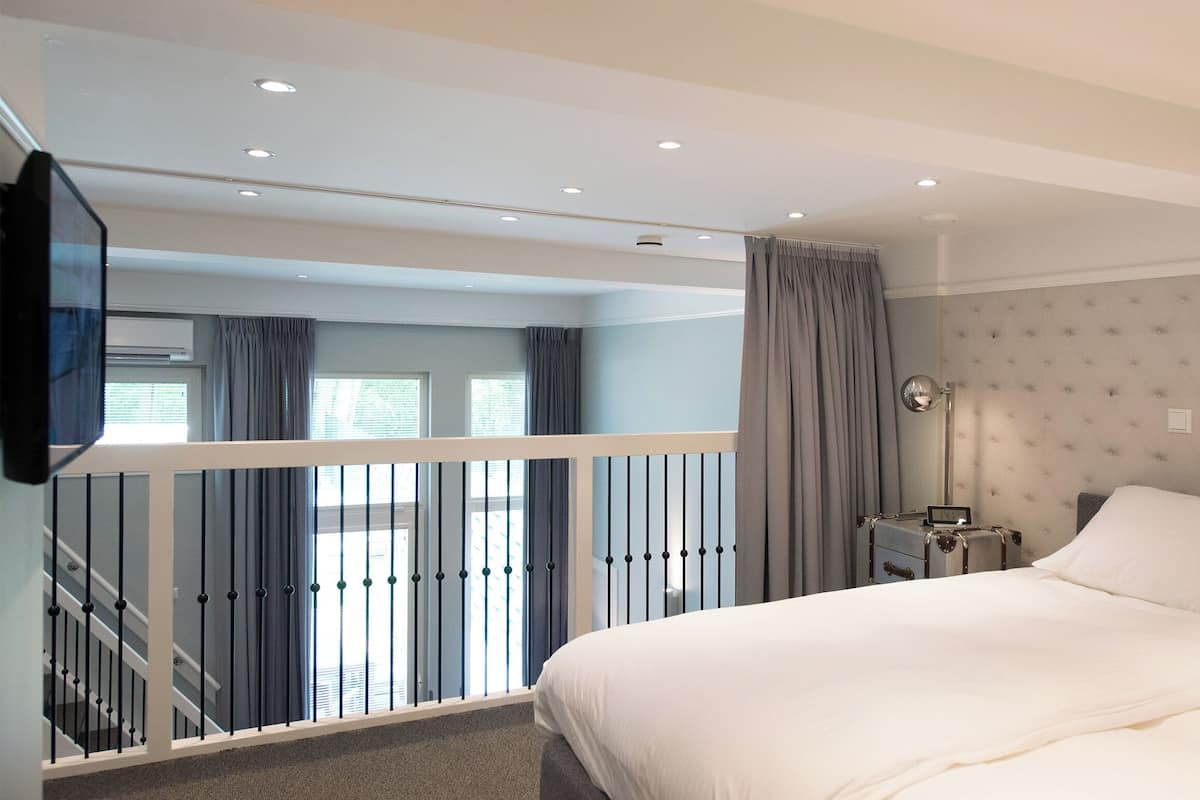 A bedroom with a bed, a nightstand, a clock, a window with curtains, a staircase with a railing, and an air conditioning unit.