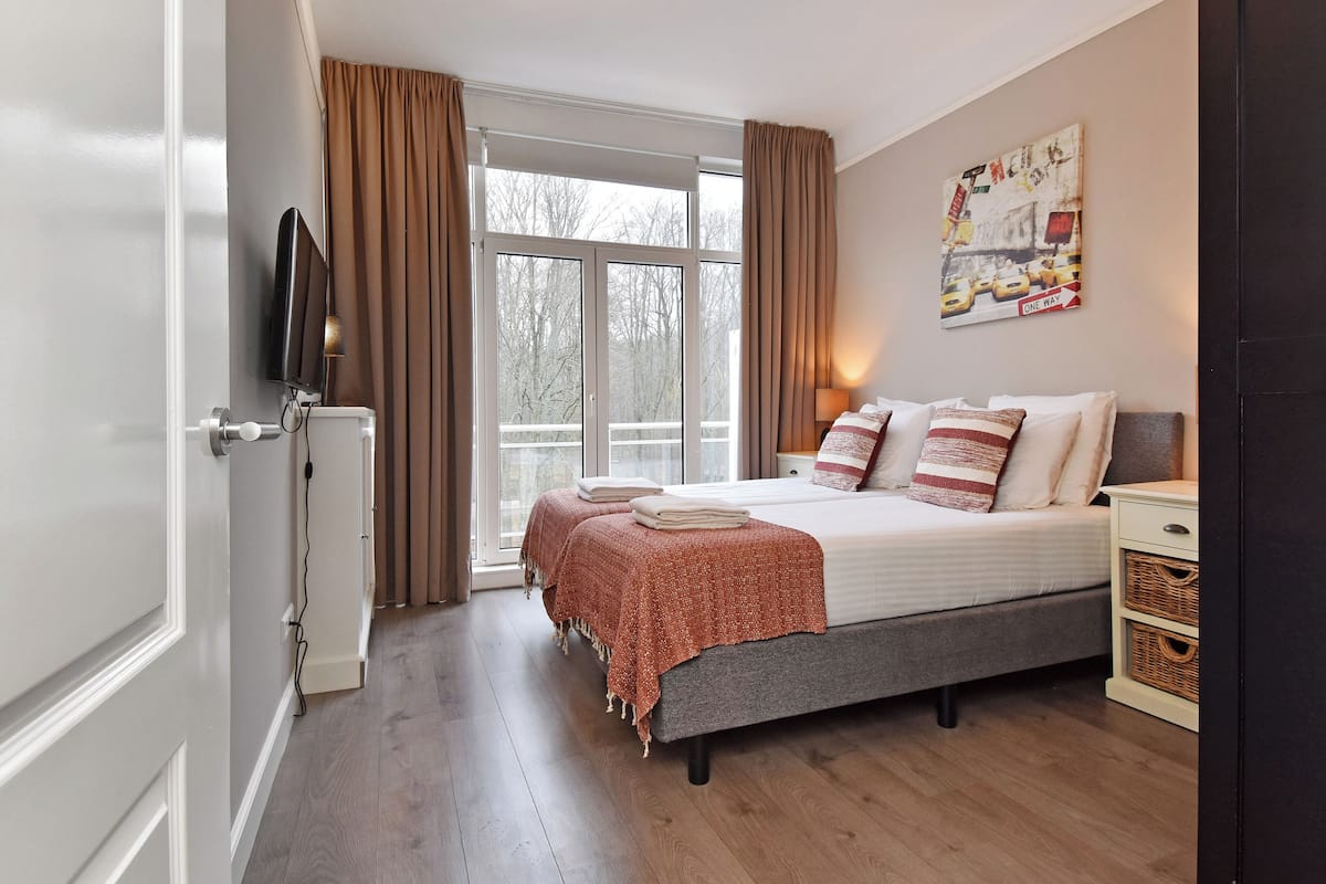 A bedroom with a bed, a TV, a window with curtains, and a painting on the wall.
