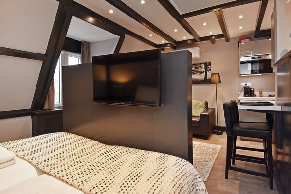 Une chambre moderne comprenant un lit, un téléviseur fixé sur un mur sombre, un bureau avec une chaise, et un coin cuisine avec une cuisinière et un réfrigérateur.
