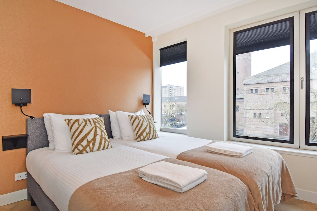 A hotel room with two beds, a large window with a view of buildings, and a wall-mounted speaker.