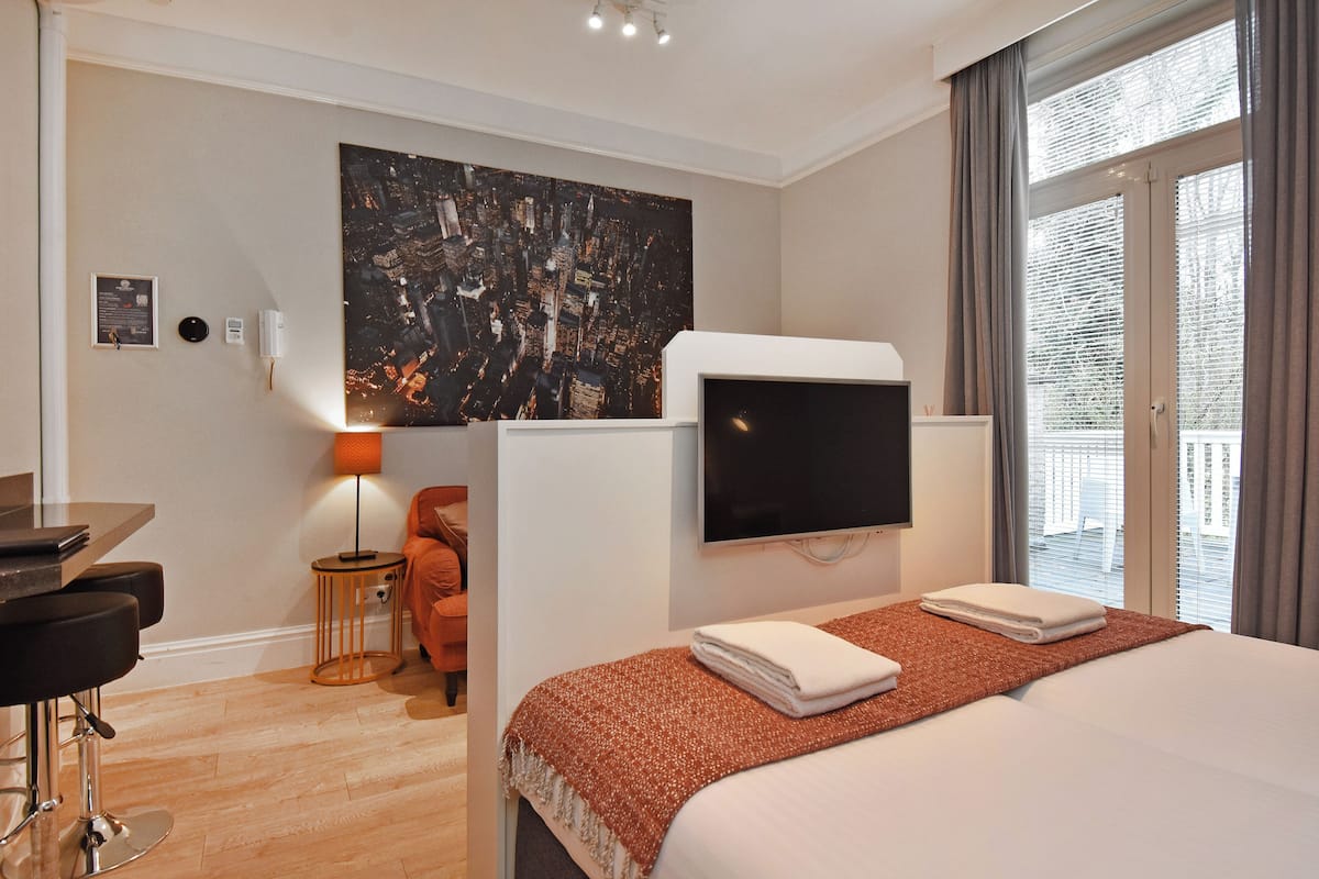 A hotel room with a bed, a TV, a desk with a chair, a small table with a lamp, and a large abstract painting on the wall.