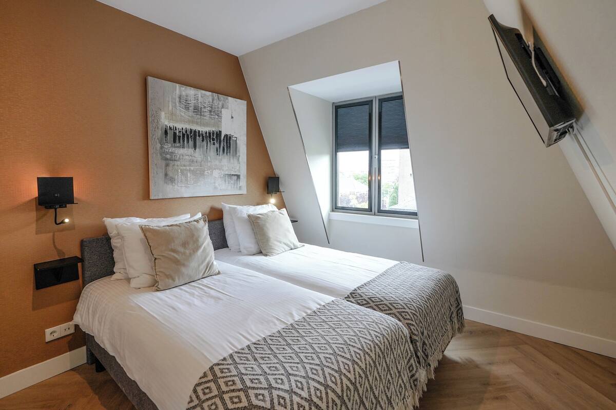 A bedroom with two beds, a large abstract painting, a window with blinds, and a mounted TV.