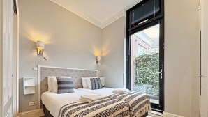 Une chambre à coucher avec un grand lit, deux lampes de chevet, une fenêtre avec des stores et une vue sur un bâtiment à l’extérieur.