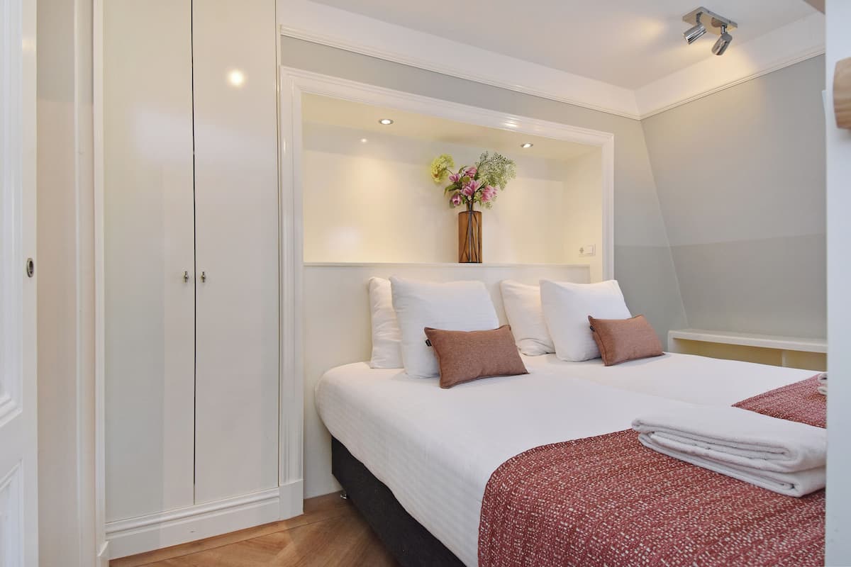 A bedroom with a large bed, white walls, a wooden wardrobe, and a mirror with a vase of flowers.