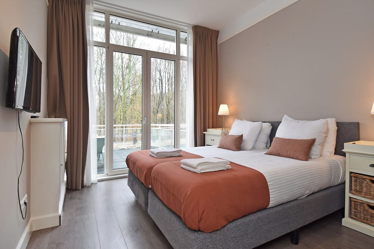 A bedroom with a large bed, a television, a nightstand, a lamp, and a view of trees through the window.