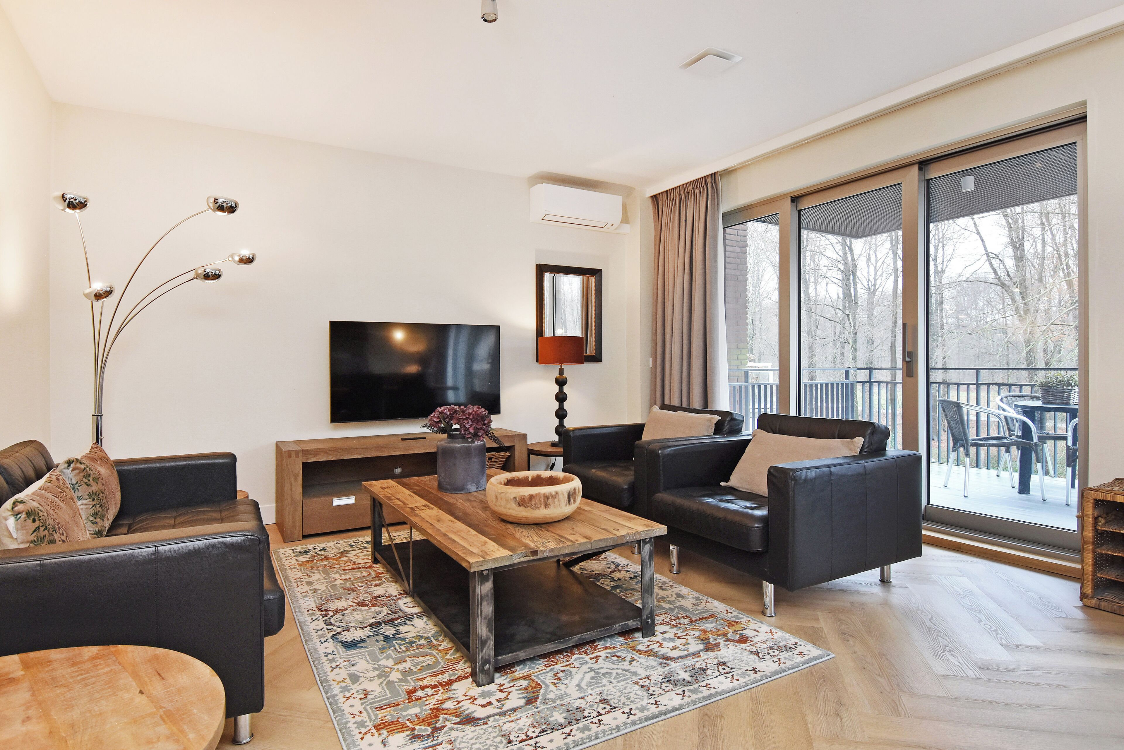 A modern living room with a flat-screen TV, two sofas, a coffee table, and a large window with a view of trees.