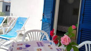 Un espace extérieur ensoleillé, aménagé avec des chaises blanches et une table, des volets bleus et un vase rempli de fleurs roses.