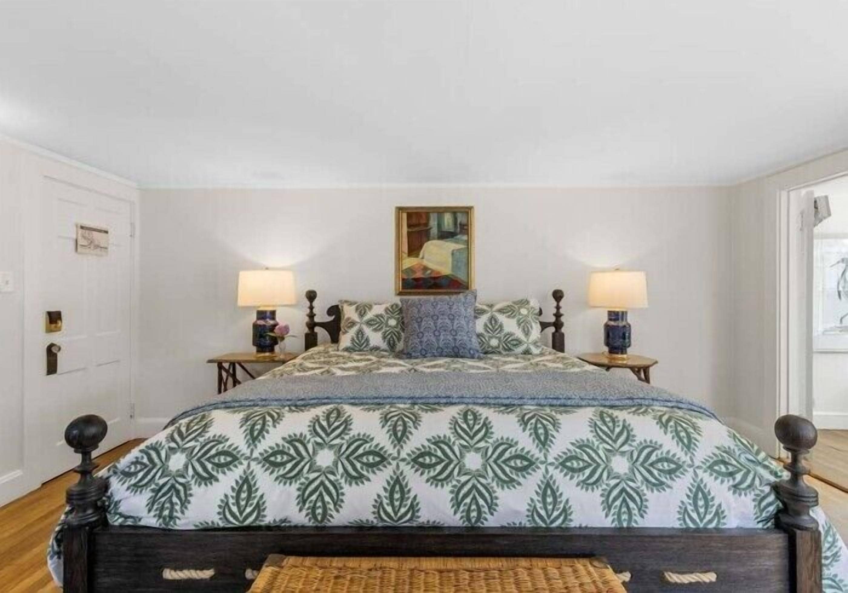 A bed with a patterned bedspread, two bedside lamps, a painting above the bed, and a door to the left.
