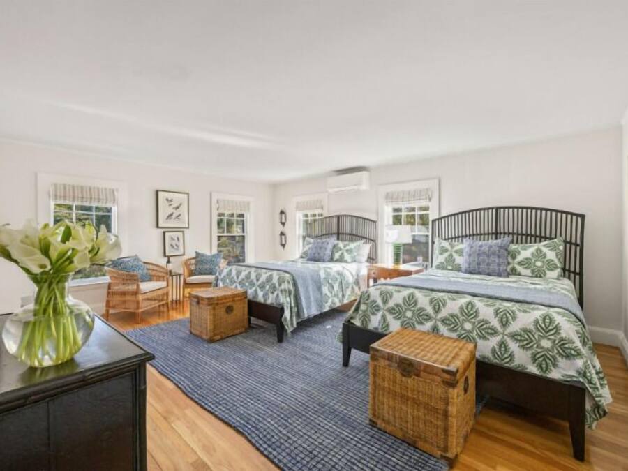 A bedroom with two beds, a desk, wicker furniture, and a vase with flowers.