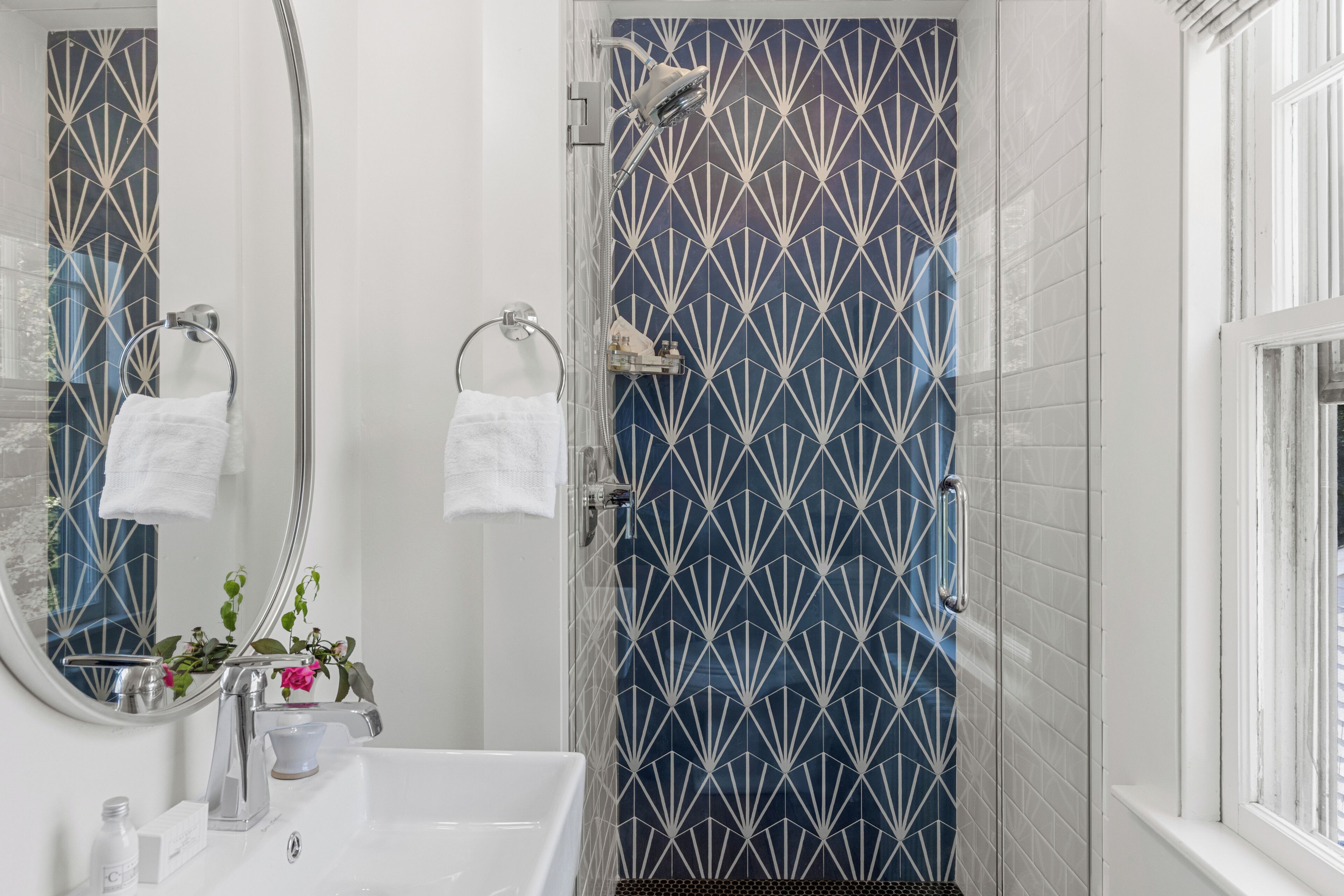 Une salle de bain avec un lavabo blanc, un miroir, une douche dont les carreaux présentent un motif géométrique et une fenêtre.
