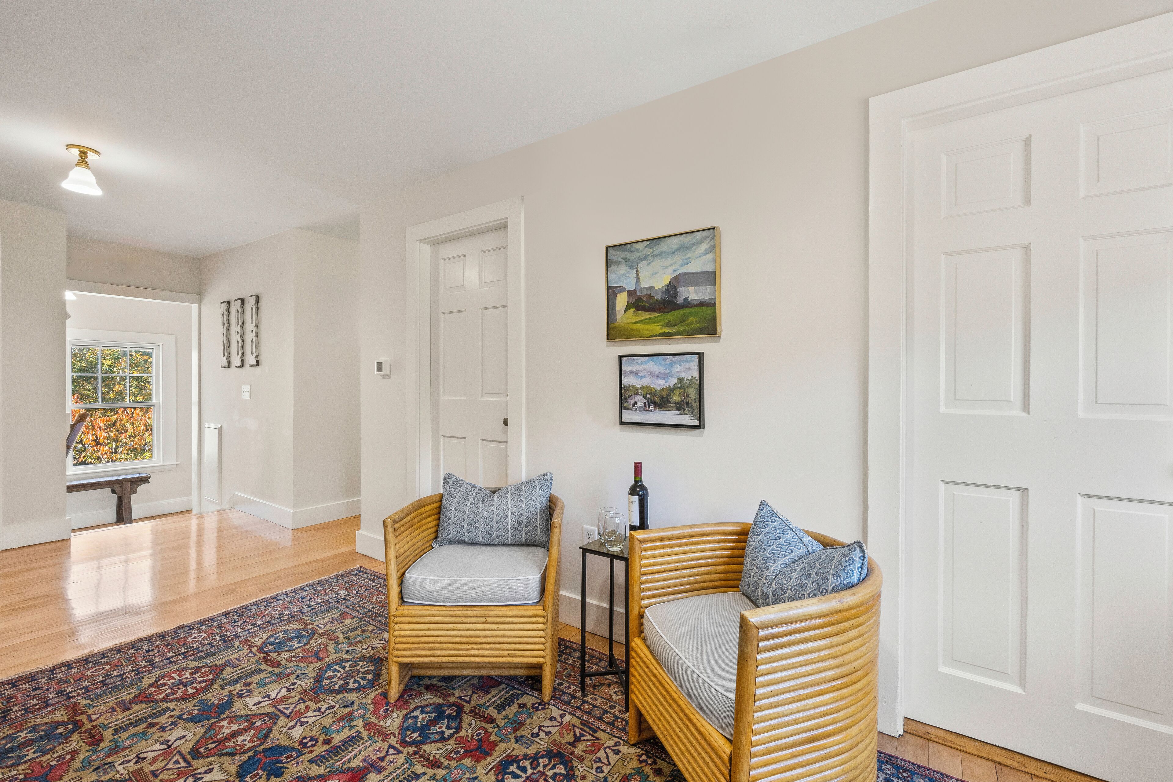 A hallway with two wicker chairs, a patterned rug, and two framed pictures on the wall.