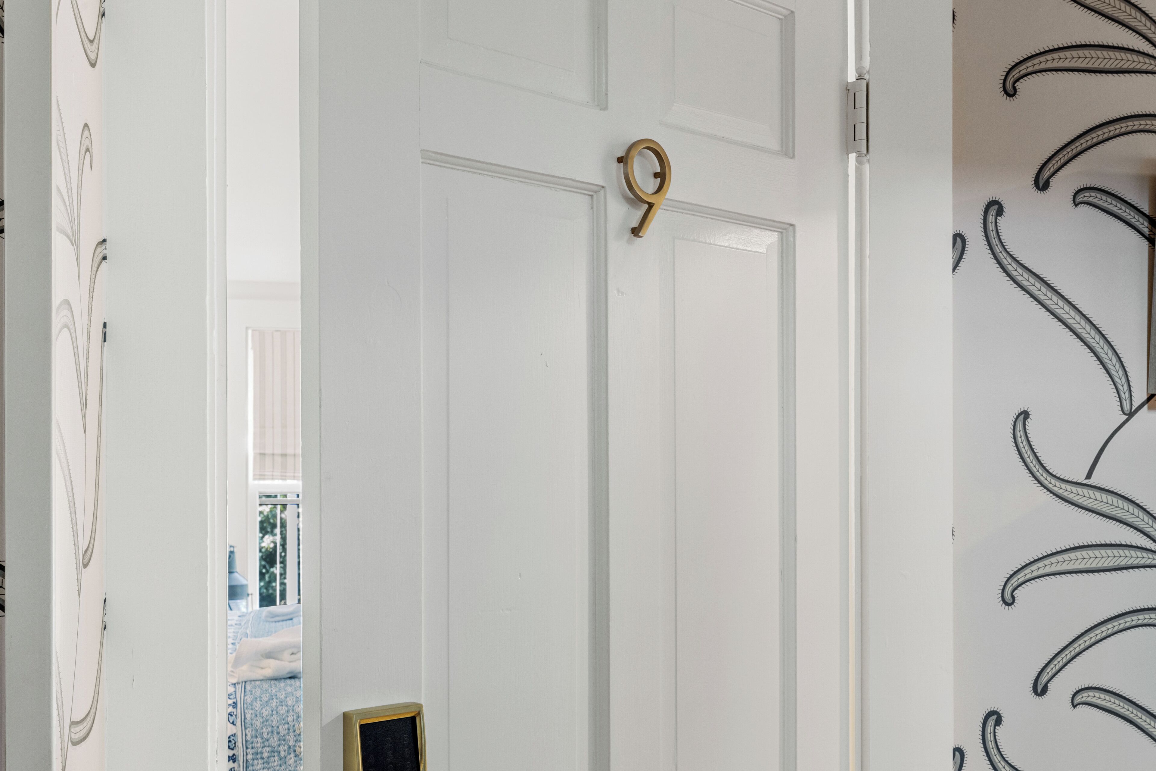 A white door with a brass handle, a patterned wall, and a glimpse of a room with a bed.