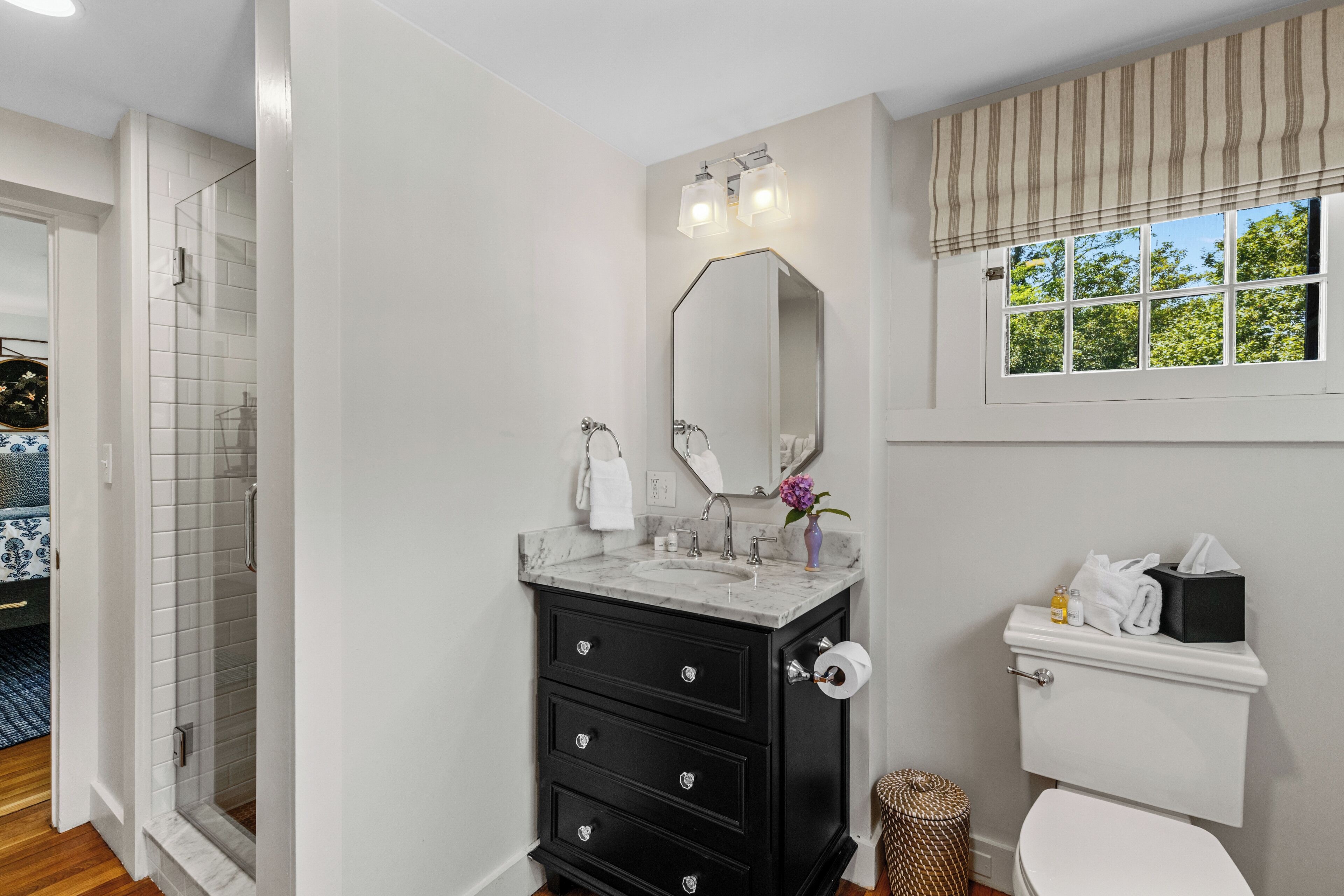 Un baño con ducha, inodoro, lavabo con espejo y ventana con cortinas.