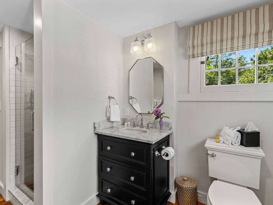 Un baño con ducha, inodoro, lavabo con espejo y ventana con cortinas.