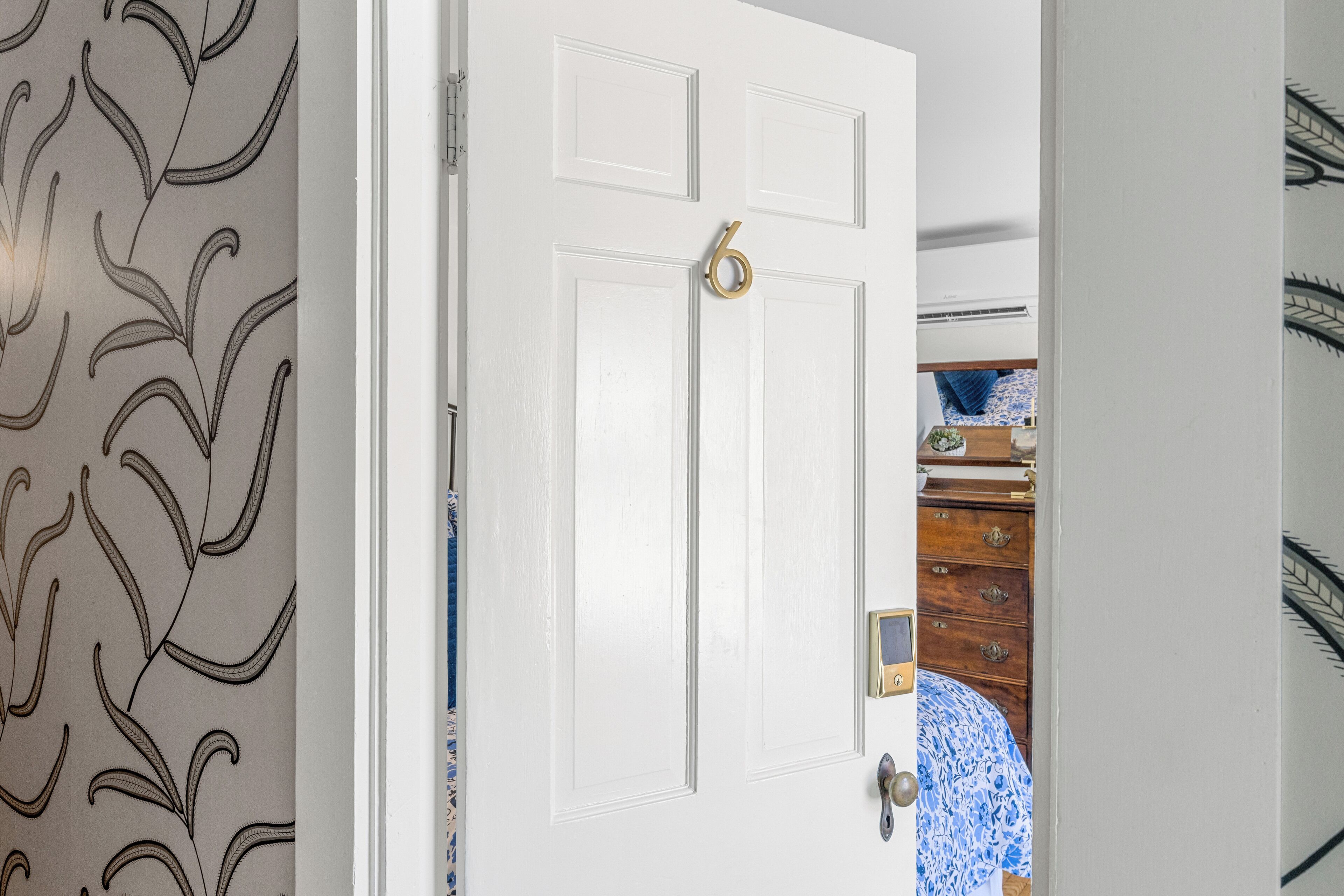 A white door with a brass handle and a keyhole, leading to a room with patterned wallpaper and a blue and white patterned bedspread.