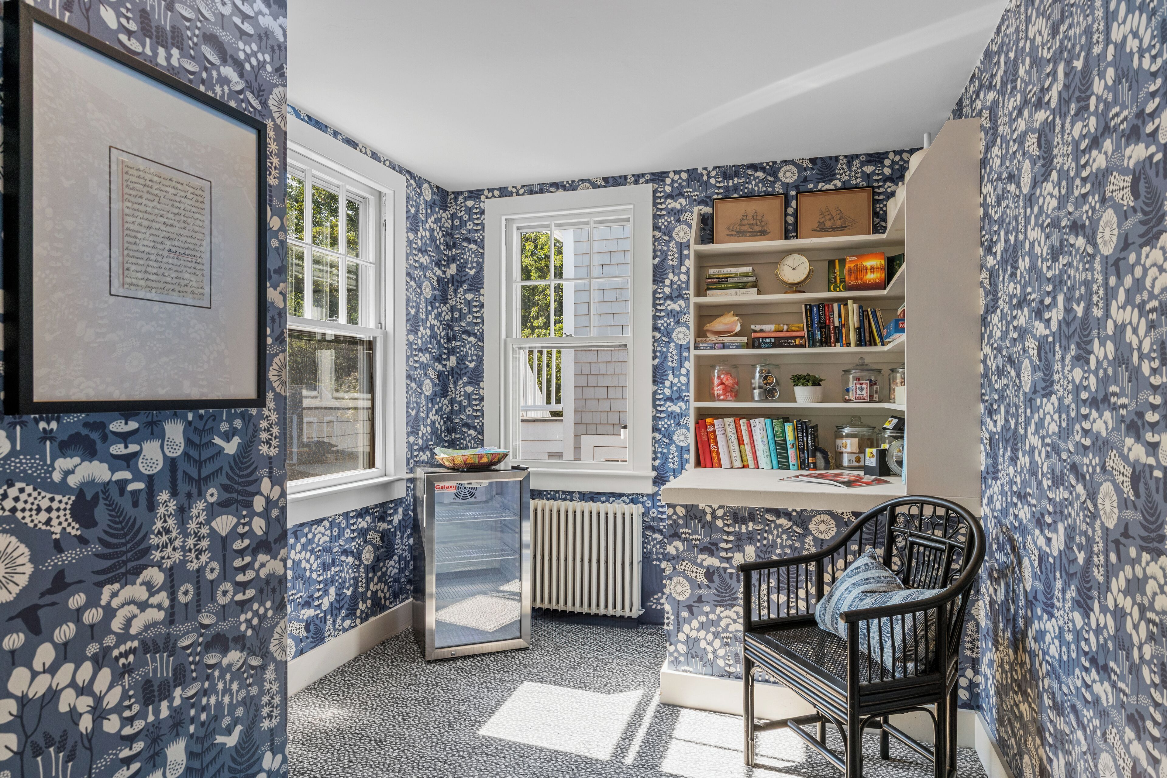 Una habitación con papel tapiz floral azul, una estantería, un radiador y una ventana con contraventanas blancas.