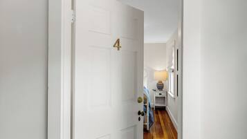 A white door with a number 4, a wooden floor, a small bedside table with a lamp, and a framed picture on the wall.