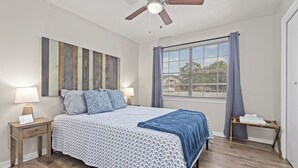 A bedroom with a bed, nightstands, a ceiling fan, a window with curtains, and a view of the outside.