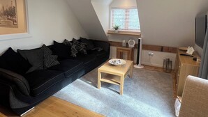 A modern living room with a black sofa, a wooden coffee table, and a small window with blinds. - Charming Countryside Barn Retreat – Perfect for Relaxing Escapes (East Preston)