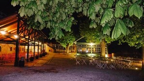 Un espacio de estar techado al aire libre con mesas y sillas, un edificio con amplios ventanales y un árbol que da sombra.