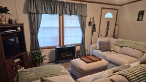 Una sala de estar con sofá, reposapiés, mueble para televisor y ventana con cortinas.