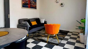 Ein modernes Wohnzimmer mit grauem Sofa, rundem Tisch, leuchtend orangefarbenem Sessel, schwarz-weiß kariertem Boden, einem Stadtbildgemälde und sechseckigen Wanddekorationen.
