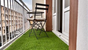 Ein Balkon mit Kunstgrasanlage, einem Klappstuhl und Blick auf ein Gebäude im Hintergrund.