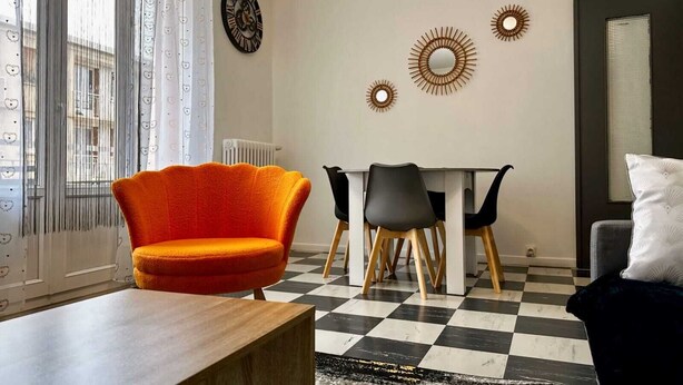 Ein modernes Wohnzimmer mit Schachbrettmuster-Boden, einem Holztisch, einem leuchtend orangefarbenen Sessel, einem Esstisch mit vier Stühlen und Wandbildern.