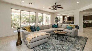 A spacious living room with a large L-shaped sectional sofa, a coffee table, and a patterned rug. There are large windows providing natural light and a view of the outdoors. - Amazing Litchfield Estate With Backyard Oasis! (Litchfield Park)