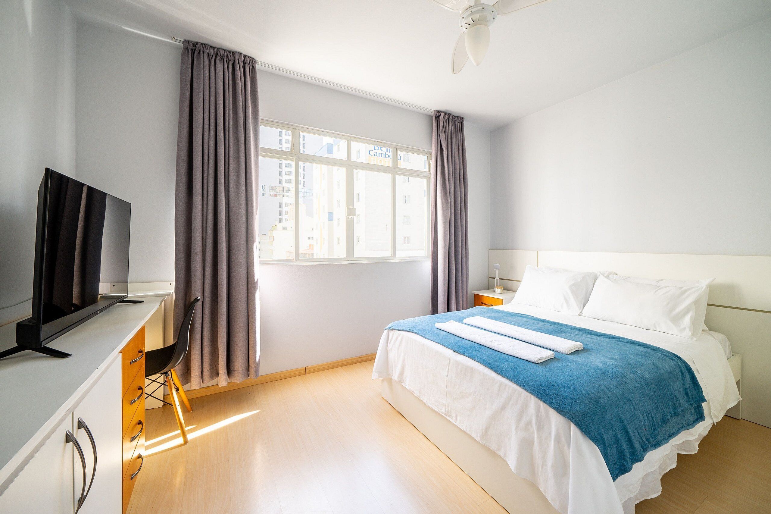 Quarto de hotel com uma cama, uma escrivaninha com televisão, uma cadeira e uma janela com cortinas.