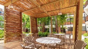 Un patio al aire libre con mesas redondas y sillas de mimbre, rodeado por una pérgola de bambú y follaje.