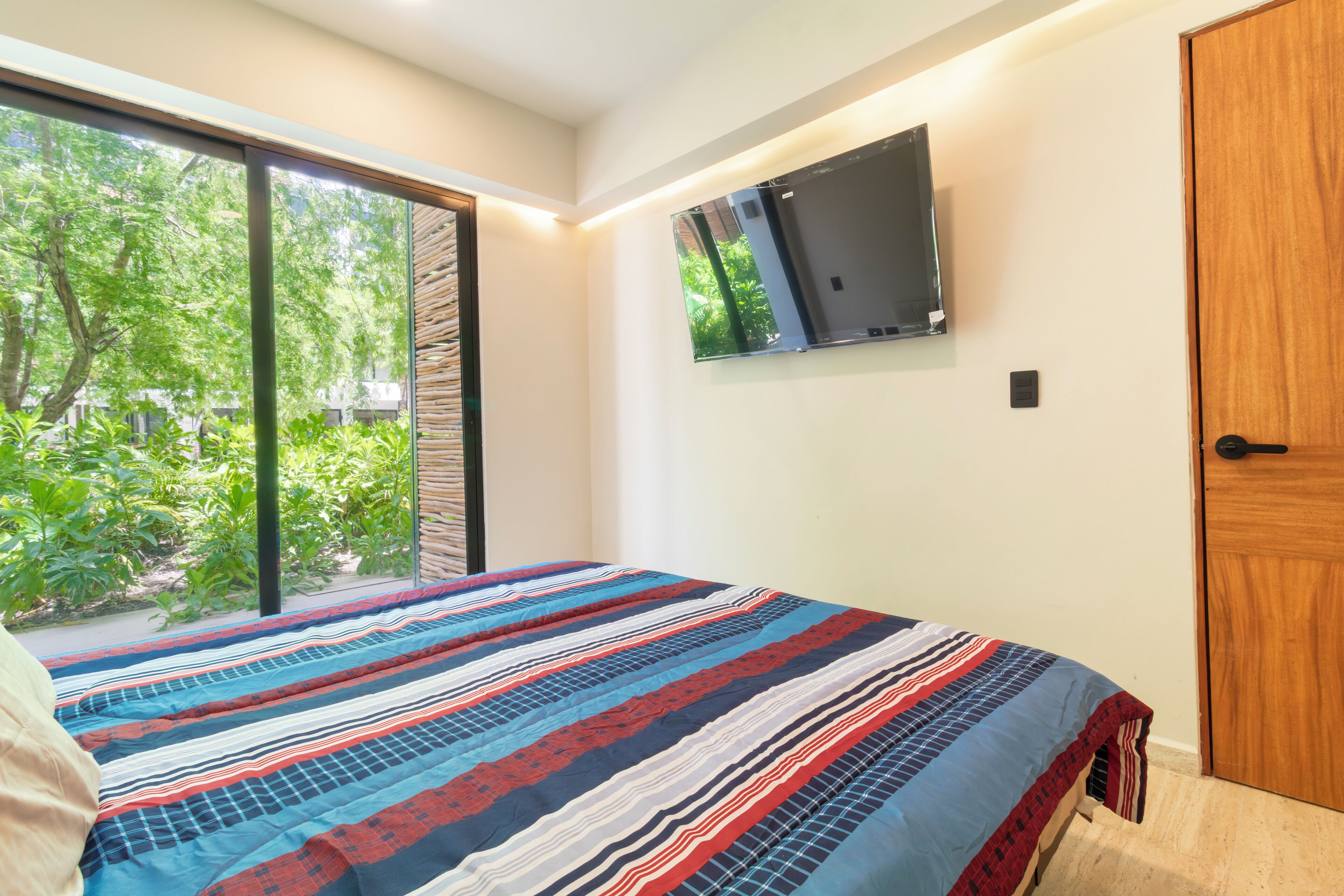 A bedroom with a large window, a bed with a striped bedspread, a mounted TV, and a wooden door.