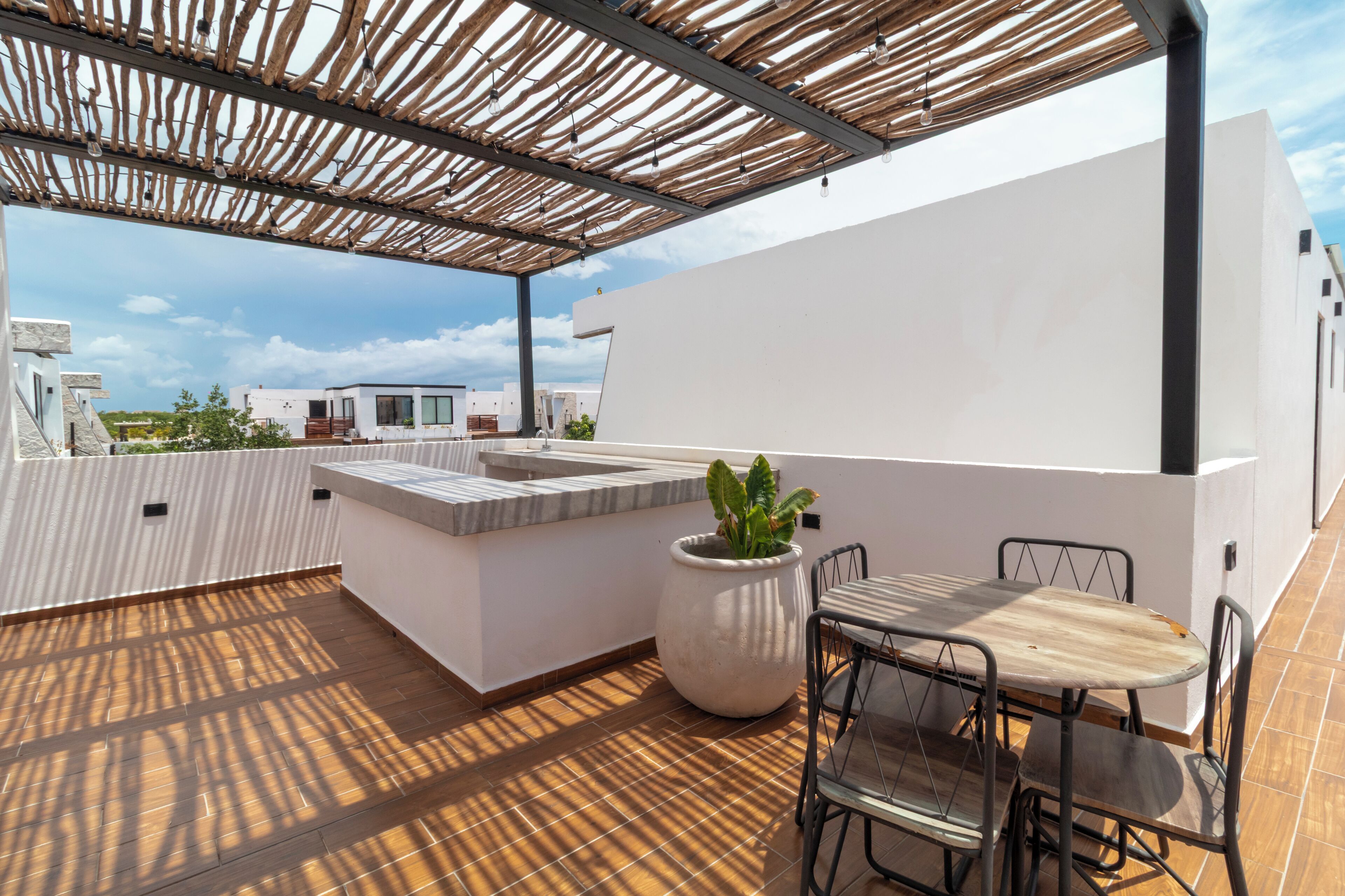 Een terras op het dak met een houten pergola, een zithoek met metalen stoelen, een grote plant in pot en een witte muur.